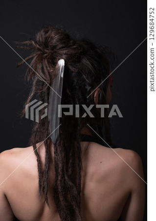 Studio portrait of woman with long dreadlocks and big feather in her hair standing back to camera Studio portrait of woman with long dreadlocks and big feather in her hair standing back to camera 129684752