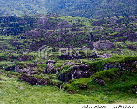 A grassy slope with massive stones is seen in this view of the Gap of Dunloe. The picturesque landscape shows vibrant green grass and rocky terrain. A grassy slope with massive stones is seen in this view of the Gap of Dunloe. The picturesque landscape shows vibrant green grass and rocky terrain. 129686166
