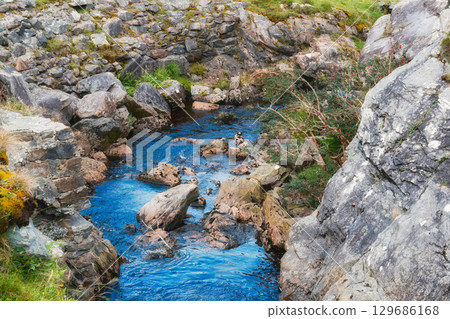 Clear blue river cascades through a narrow gorge filled with various sized rocks. Green foliage and moss cling to the rock walls surrounding the river at Gap of Dunloe. 129686168