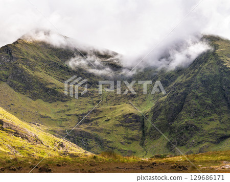 Carrauntoohill peak, the highest point in Ireland, is partially covered by atmospheric clouds. Green hillsides and sparse vegetation are seen along the popular Carrauntoohill walkway. 129686171