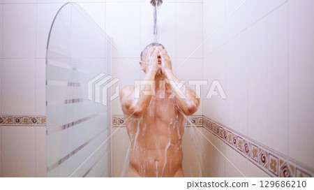 Man washing face and body while taking shower in bathroom 129686210