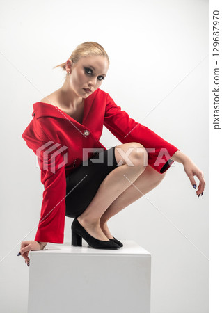 Beautiful woman with red jacket and mini shirt sitting on white cube 129687970