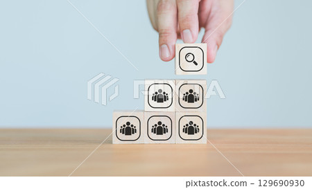 Hand placing a magnifying glass icon wooden cube block above people group wood blocks, representing talent search, company, leadership, recruitment, or leadership selection strategy. 129690930