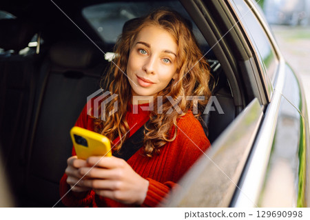 Positive young woman in casual clothes sits in the back seat of a car with a mobile phone. 129690998