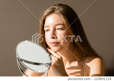 Young woman reflected in the mirror in soft evening light in a cozy room. Beauty and care concept. 129691124