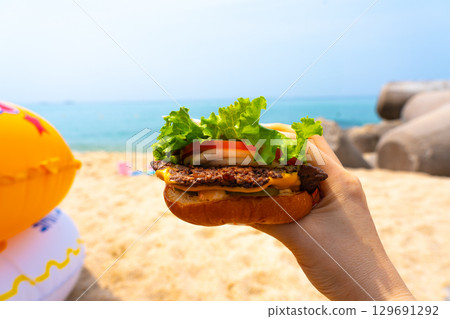 Woman holding burger in a hand against beach background 129691292