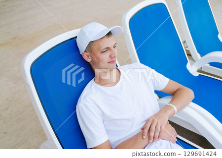 Man wearing a white shirt and a white hat is sitting on a blue chair 129691424