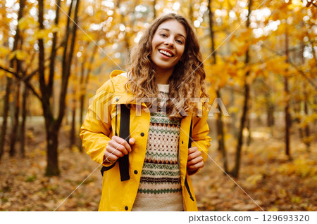 Hiking in autumn forest. Smiling tourist in bright coat walks through autumn park in sunny weather. 129693320