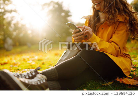 A smiling woman sits outdoors on the lawn among yellow autumn leaves with a phone in her hands. 129694120