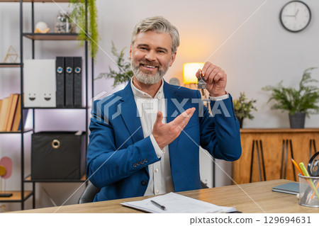 Middle-aged businessman signs lease holds apartment keys smiles shakes agent hand marking milestone 129694631