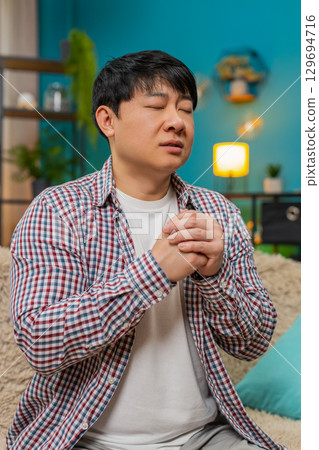 Asian man with eyes closed repeating mantra, calm meditative prayer, hopeful quiet spiritual moment 129694716
