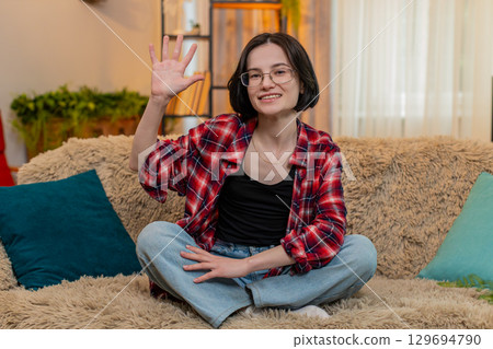 Young woman smiling and waving hand at home, greeting someone with friendly and warm expression Young woman smiling and waving hand at home, greeting someone with friendly and warm expression 129694790