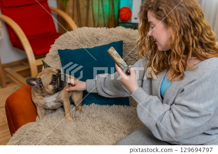 Young woman taking photo of cute pug dog on home sofa, smiling and creating cheerful pet content Young woman taking photo of cute pug dog on home sofa, smiling and creating cheerful pet content 129694797