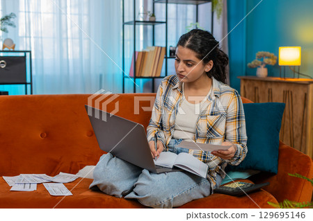 Indian woman working on laptop calculating family budget writing notes using calculator and receipts 129695146