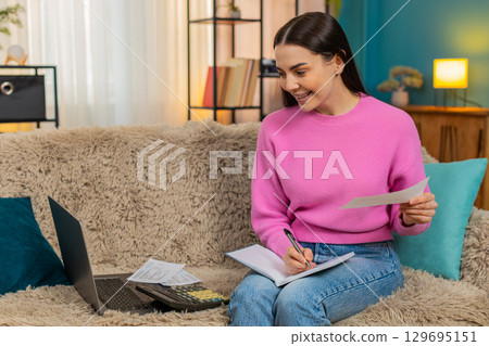 Young woman on sofa with laptop and calculator counting expenses worried writing notes in notebook Young woman on sofa with laptop and calculator counting expenses worried writing notes in notebook 129695151