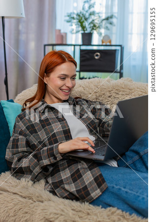 Woman sitting on sofa shopping online with laptop choosing dress touching chin in thoughtful pose 129695155