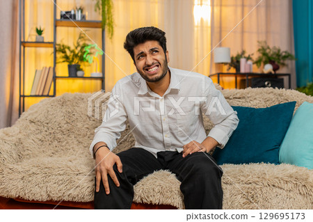 Indian young man in pain holding injured knee grimacing suffering from leg injury feeling discomfort Indian young man in pain holding injured knee grimacing suffering from leg injury feeling discomfort 129695173