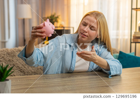 Young woman shaking empty piggy bank at home, frustration, no coins left, disappointed money check 129695196