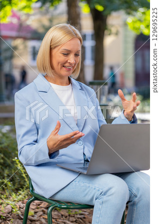 Mature businesswoman talking on video call using laptop while sitting on bench in city street 129695225