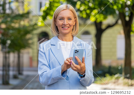 Smiling mature Caucasian businesswoman in suit using smartphone and looking at camera on city street Smiling mature Caucasian businesswoman in suit using smartphone and looking at camera on city street 129695232
