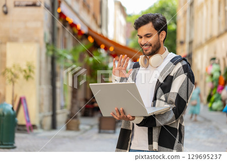 Young Indian man freelancer sitting on street, looking at laptop camera, making video call outdoors 129695237