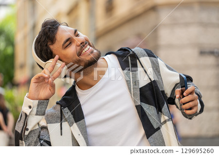 Happy Indian man wearing headphones choosing, listening disco rock n roll music dancing on street 129695260