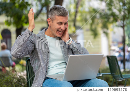 Happy man freelancer working on laptop screams in triumph win celebrating success sitting on chair 129695278