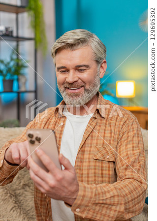 Mature man sitting on sofa with smartphone browsing photos, smiling and liking favorites at home Mature man sitting on sofa with smartphone browsing photos, smiling and liking favorites at home 129695302