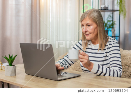 Mature old woman working from home on laptop, writing report, replying emails, sipping coffee Mature old woman working from home on laptop, writing report, replying emails, sipping coffee 129695304