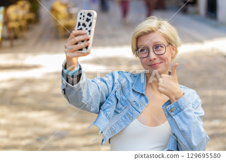 Cheerful young woman smiling friendly at camera waving hands gesturing while standing on city street 129695580