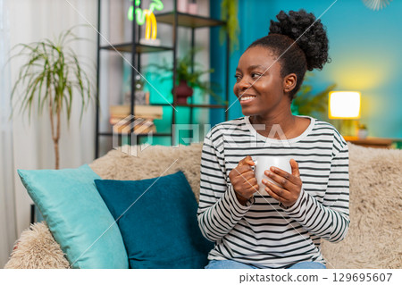 African American young woman relaxing at home on couch, holding hot tea cup, dreaming, smiling African American young woman relaxing at home on couch, holding hot tea cup, dreaming, smiling 129695607