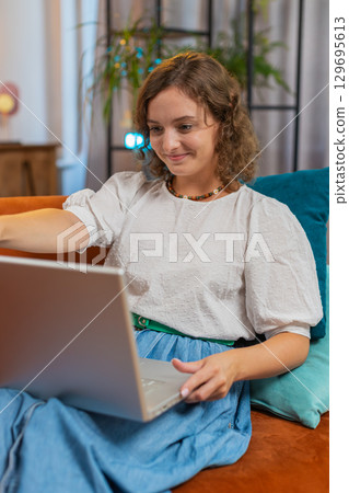 Happy young woman lady sitting on sofa opens laptop and working on project in living room at home 129695613