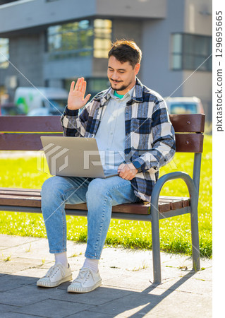 Mature businessman on video call using laptop while sitting on bench in downtown city street Mature businessman on video call using laptop while sitting on bench in downtown city street 129695665