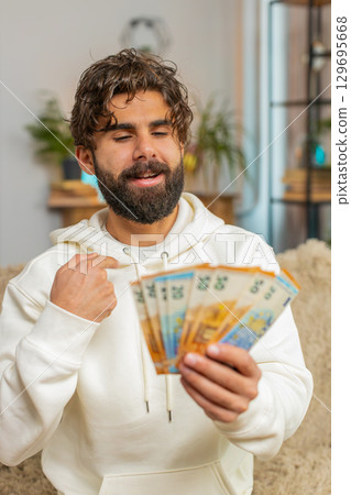 Successful rich young Hispanic man holding euro bill money fan sitting on sofa couch at home 129695668