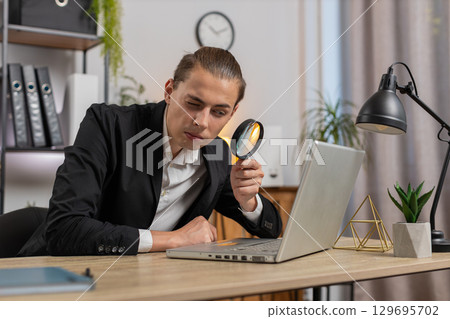 Surprised researcher businessman working at home office holding magnifying glass looking at laptop 129695702