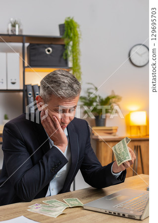 Upset businessman freelancer counting dollar cash insufficient amount of money at home office desk 129695703