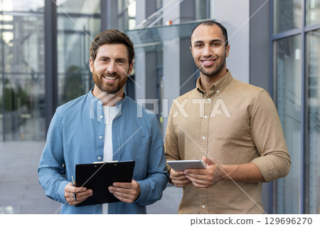 Two men stand outside a modern office building. They hold a clipboard and tablet, smiling professionally, representing teamwork or professional collaboration in a business environment. 129696270