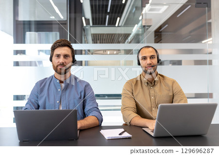 Two professionals wearing headsets work on laptops at a desk in office. The setting suggests customer service or technical support, reflecting collaboration, and a professional working environment. 129696287