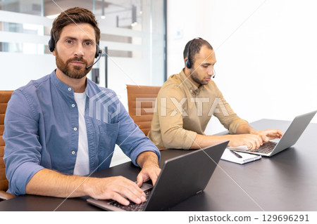 Two office professionals using laptops, wearing headsets, and engaged in a focused environment. They collaborate in an office setting, symbolizing communication, teamwork, and technology-driven 129696291