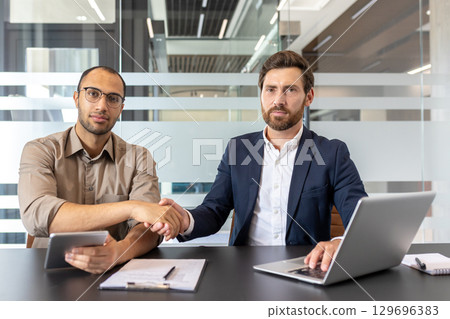 Business professionals sitting in an office setting, shaking hands to finalize a collaboration. They are equipped with a laptop and notes showcasing teamwork and agreement in a managerial environment Business professionals sitting in an office setting, shaking hands to finalize a collaboration. They are equipped with a laptop and notes showcasing teamwork and agreement in a managerial environment 129696383