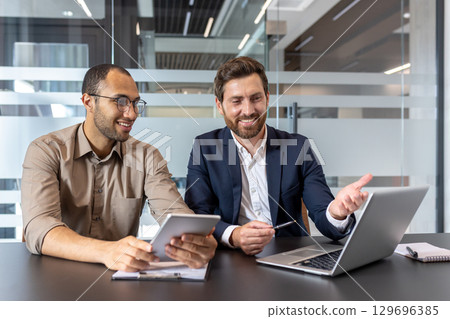 Business colleagues working together on a laptop and tablet, collaborating on a project in a well-lit, modern office environment. Business colleagues working together on a laptop and tablet, collaborating on a project in a well-lit, modern office environment. 129696385