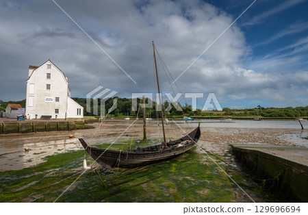 Old wooden boat with mast sitting on mud. Old tide mill behind. View of the quay at low tide in the River Deben at the historic town of Woodbridge in Suffolk, UK. Old wooden boat with mast sitting on mud. Old tide mill behind. View of the quay at low tide in the River Deben at the historic town of Woodbridge in Suffolk, UK. 129696694
