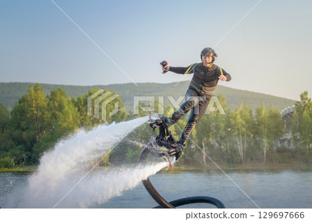 A flyboarding athlete soars above a tranquil lake during the golden hour, showcasing remarkable skills and gravity-defying maneuvers. The backdrop features lush green trees and distant hills, 129697666