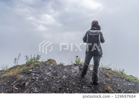 A person stands at the edge of a rocky hillside, gazing into the distance as low clouds create a misty atmosphere. The early morning light pierces through the gray sky, illuminating the damp terrain. 129697684