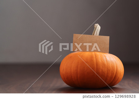 Small orange pumpkin on black walnut table with white wall background and empty paper card 129698928