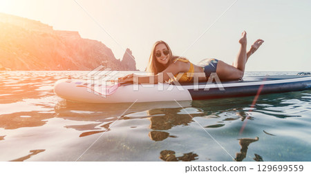 Woman Paddleboarding Ocean Coastline - A young woman lies on a paddleboard in the ocean, enjoying a day of leisure, with a scenic coastline in the background. Woman Paddleboarding Ocean Coastline - A young woman lies on a paddleboard in the ocean, enjoying a day of leisure, with a scenic coastline in the background. 129699559