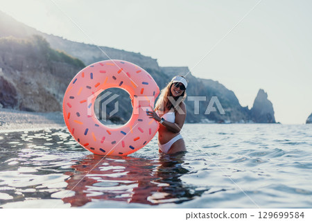 Woman, donut, ocean. Happy young woman holds inflatable donut in the sea near a rocky beach on summer vacation 129699584