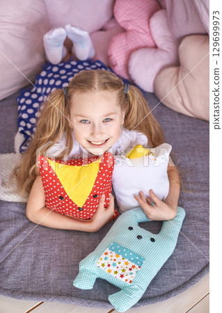 Smiling caucasian young girl playing with colorful stuffed toys in a cozy pillow 129699973