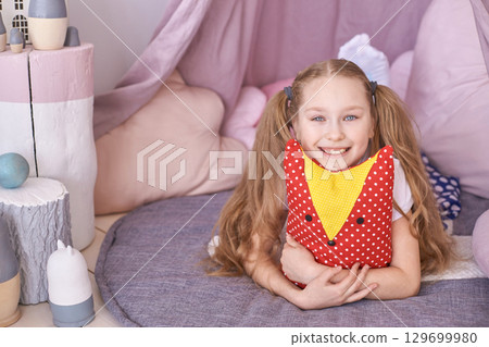 Happy caucasian young girl in cozy playroom holding colorful pillow 129699980