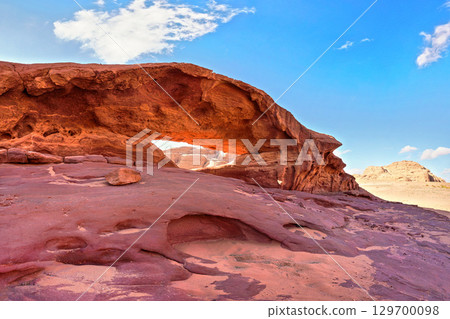Little arc or small rock window formation in Wadi Rum desert, bright sun shines on red dust and rocks, blue sky above 129700098